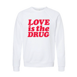 White crewneck sweatshirt with bold bright orangey red 'Love is the Drug' text across the chest